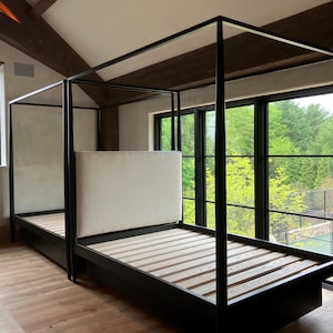 May include: A modern black four-poster bed with a light-colored headboard and wooden slats. The bed frame is set against a backdrop of large windows overlooking a lush green landscape. The room features hardwood floors and exposed wooden beams.
