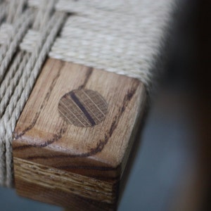 May include: Close-up of a wooden chair with a woven rope seat. The chair has a natural wood finish and a round wooden dowel at the corner.