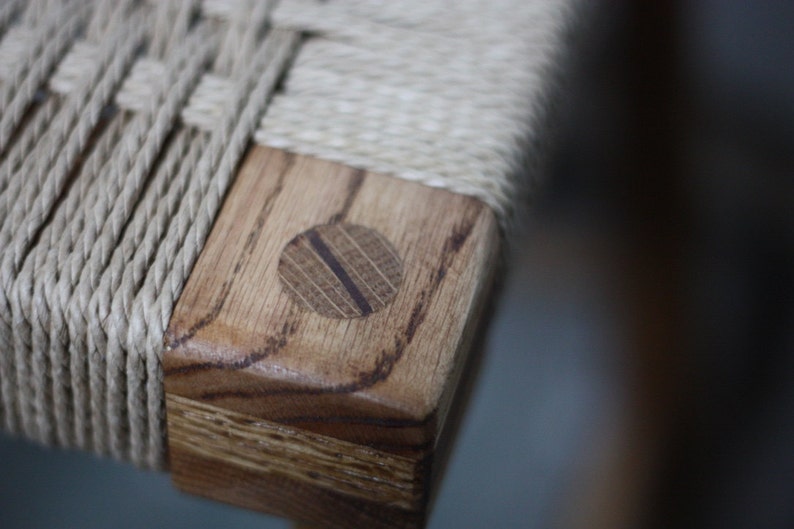 May include: Close-up of a wooden chair with a woven rope seat. The chair has a natural wood finish and a round wooden dowel at the corner.