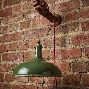 May include: A wall-mounted lamp featuring a dark brown wooden articulated hand extending from a circular base. A red fabric-covered cord hangs from the hand, supporting a glossy, dark green metal dome-shaped shade with a brass-colored light socket.