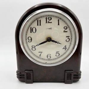 May include: A vintage brown clock with a cream-coloured face and black numerals. The clock has a silver-coloured bezel and black hands. The clock's case is a dark brown colour with a rounded top and rectangular base. The clock measures approximately 18 cm in height.