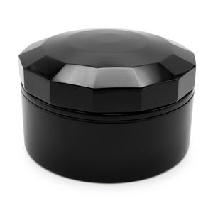 May include: A black, round container with a faceted lid. The lid's design resembles a gemstone, with multiple flat surfaces reflecting light. The container's base is a smooth, cylindrical shape. The overall aesthetic is sleek and modern.