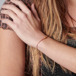 Puede incluir: Un brazalete de cuerda roja con un pequeño dije de oro. El dije es una simple forma ovalada.