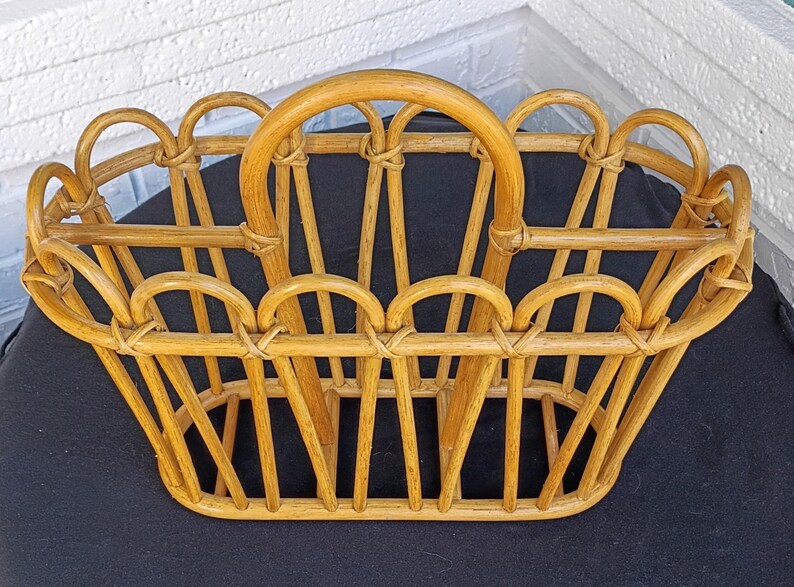 Puede incluir: Una cesta de rat&aacute;n tejido de color marr&oacute;n claro con un asa. La cesta tiene una base rectangular y una serie de aberturas arqueadas a los lados. El asa es una pieza grande y curvada de rat&aacute;n. La cesta est&aacute; dise&ntilde;ada para transportar objetos.