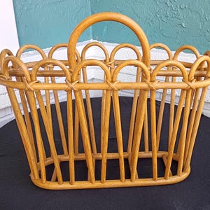 Puede incluir: Una cesta de rat&aacute;n tejido de color marr&oacute;n claro con un asa curva. La cesta tiene una base rectangular y soportes verticales, con detalles arqueados decorativos a lo largo del borde superior. La cesta est&aacute; dise&ntilde;ada para transportar objetos.