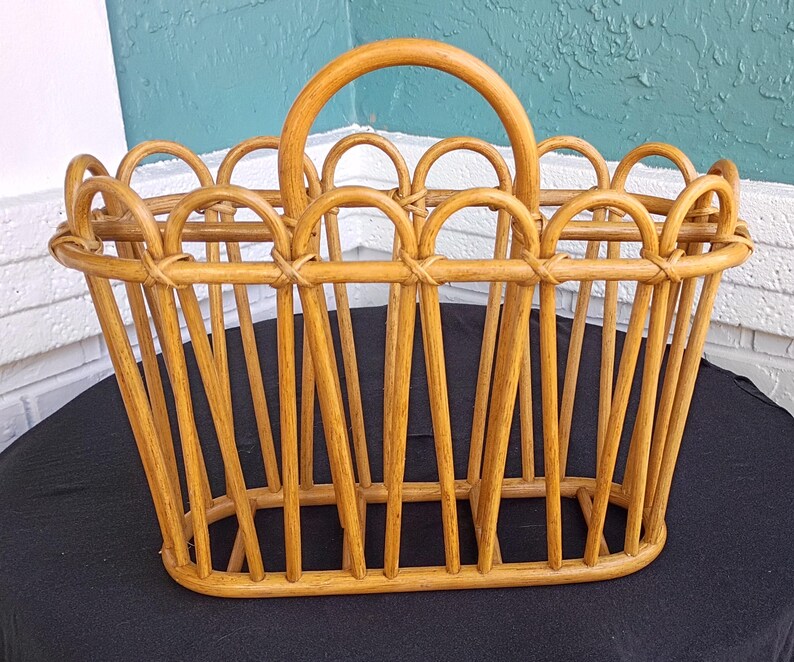 Puede incluir: Una cesta de rat&aacute;n tejido de color marr&oacute;n claro con un asa curva. La cesta tiene una base rectangular y soportes verticales, con detalles arqueados decorativos a lo largo del borde superior. La cesta est&aacute; dise&ntilde;ada para transportar objetos.