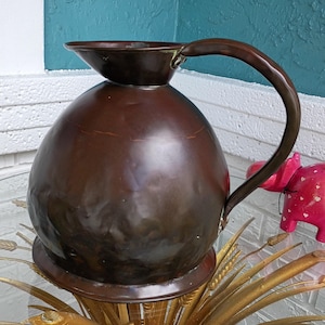 May include: A vintage copper pitcher with a rounded body and a curved handle. The pitcher has a spout and sits on a decorative base. The copper has a rich, dark brown patina, suggesting age and use. The pitcher is likely used for serving beverages.