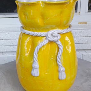 May include: A bright yellow ceramic vase with a white rope detail tied around the middle. The vase has a ruffled top and a rounded shape. The rope has tassels at the ends, adding a decorative touch. The vase is sitting on a gray surface.
