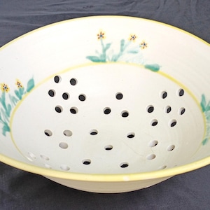 May include: A round, off-white ceramic colander featuring a yellow rim and hand-painted green and yellow floral motifs along the interior sides. The bowl has numerous circular drainage holes distributed across the bottom and lower sides.