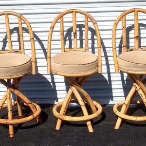 May include: Three brown bamboo bar stools with beige cushions. The stools have a twisted base and a curved back.