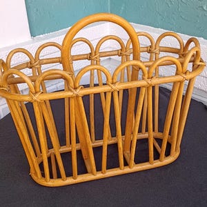 Puede incluir: Una cesta rectangular de rat&aacute;n marr&oacute;n claro con asa. La cesta tiene un borde superior festoneado y soportes verticales. El asa es arqueada y la cesta est&aacute; sobre una superficie negra.