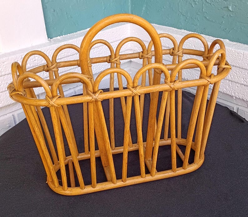 Puede incluir: Una cesta rectangular de rat&aacute;n marr&oacute;n claro con asa. La cesta tiene un borde superior festoneado y soportes verticales. El asa es arqueada y la cesta est&aacute; sobre una superficie negra.