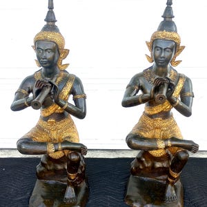 Puede incluir: Dos estatuas de bronce de figuras tocando instrumentos musicales. Las figuras están arrodilladas y visten tocados y ropa elaborados. Las estatuas están decoradas con detalles dorados.