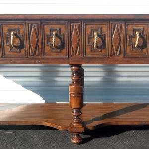 May include: A dark brown wooden console table with six drawers and a lower shelf. The drawers have carved details and rope-like handles. The table has turned legs and a curved lower shelf.