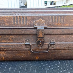 May include: A weathered, rectangular metal trunk with a hinged lid and a latch closure. The trunk is a rusty brown color, with visible signs of age and wear. It features a handle on the front and decorative vertical lines on the top.