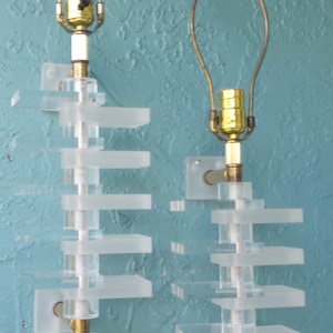 May include: A pair of white acrylic wall sconces with gold metal accents and clear glass bulbs. The sconces have a geometric design with multiple layers of rectangular shapes.