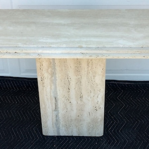 May include: A travertine console table with a rectangular top and a single pedestal base. The table is made of natural stone with a light beige color and a textured surface.