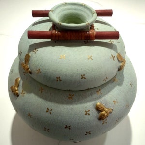 May include: A light blue ceramic vase with a stacked design and a woven bamboo handle. The vase is decorated with gold bee motifs and a pattern of small gold stars.