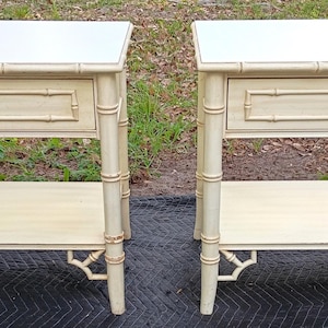 May include: Two matching cream-colored side tables with a bamboo-style design. Each table has a white top, a single drawer with a brass knob, and a lower shelf. The legs and frame have a bamboo-like appearance, adding a touch of elegance.