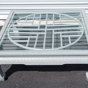 May include: A square gray coffee table with a glass top and a circular lattice design in the center. The table has four legs with a traditional Asian design.