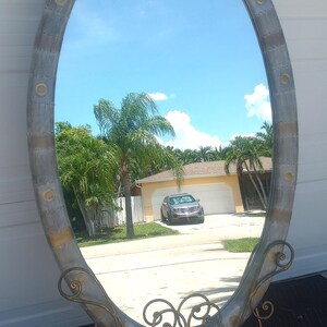 May include: Oval-shaped decorative mirror with a distressed silver-tone frame and gold accents. The frame has a riveted design with circular gold details. The mirror is supported by a gold, ornate stand. The reflection shows a blue sky and a house.