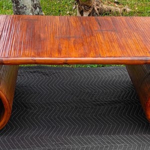 May include: A mid-century modern coffee table made of bamboo. The table has a rectangular top and two scrolled legs. The bamboo has a warm, reddish-brown finish. The table is placed on a black surface with a zigzag pattern.
