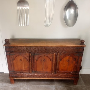 May include: A wooden chest with intricate carvings and three arched doors. The chest is a warm brown color and has decorative details along the top and bottom edges. Two decorative metal utensils hang above the chest.