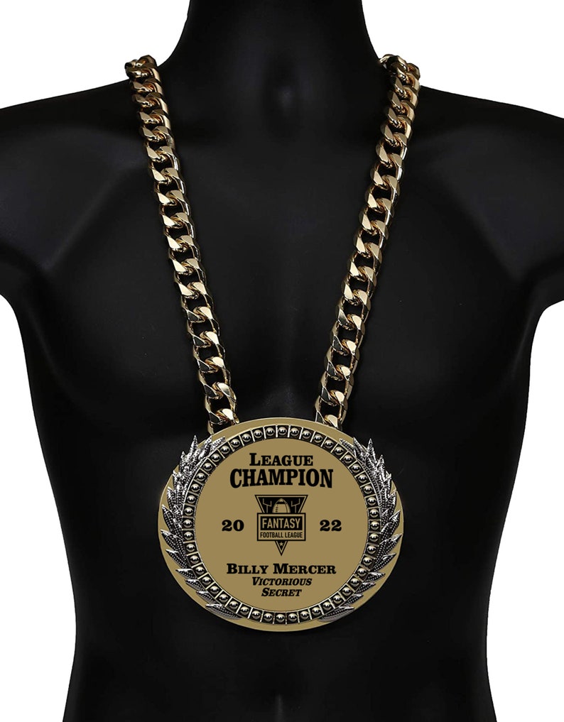 May include: A gold chain necklace with a large gold medal pendant. The medal has a black and white wreath design around the edge and says "League Champion" in black text. Inside the wreath is a black and white logo for a fantasy football league. The medal also says "20" and "22" in black text. Below the logo, it says "Billy Mercer Victorious Secret" in black text.