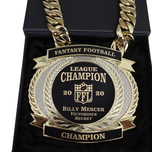 May include: A gold chain necklace with a large, round, black and gold trophy pendant. The pendant features the text "Fantasy Football League Champion 20 FFL 20 Billy Mercer Victorious Secret Champion".
