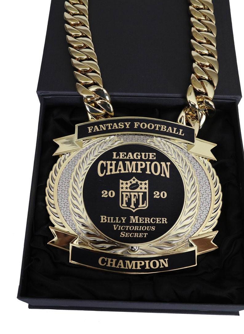 May include: A gold chain necklace with a large, round, black and gold trophy pendant. The pendant features the text "Fantasy Football League Champion 20 FFL 20 Billy Mercer Victorious Secret Champion".