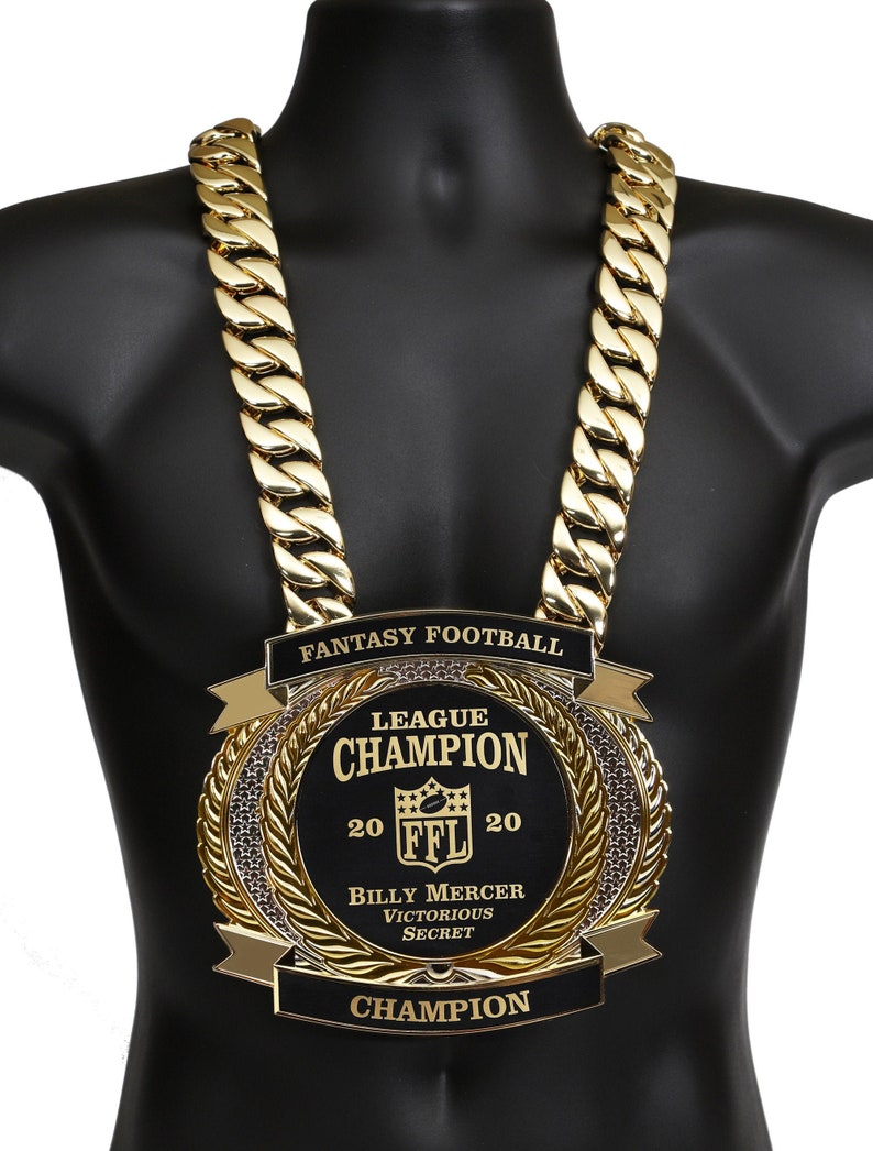 May include: A gold chain necklace with a large, black and gold fantasy football championship medal. The medal features the text "League Champion", "20 FFL 20", "Billy Mercer", "Victorious Secret", and "Champion".