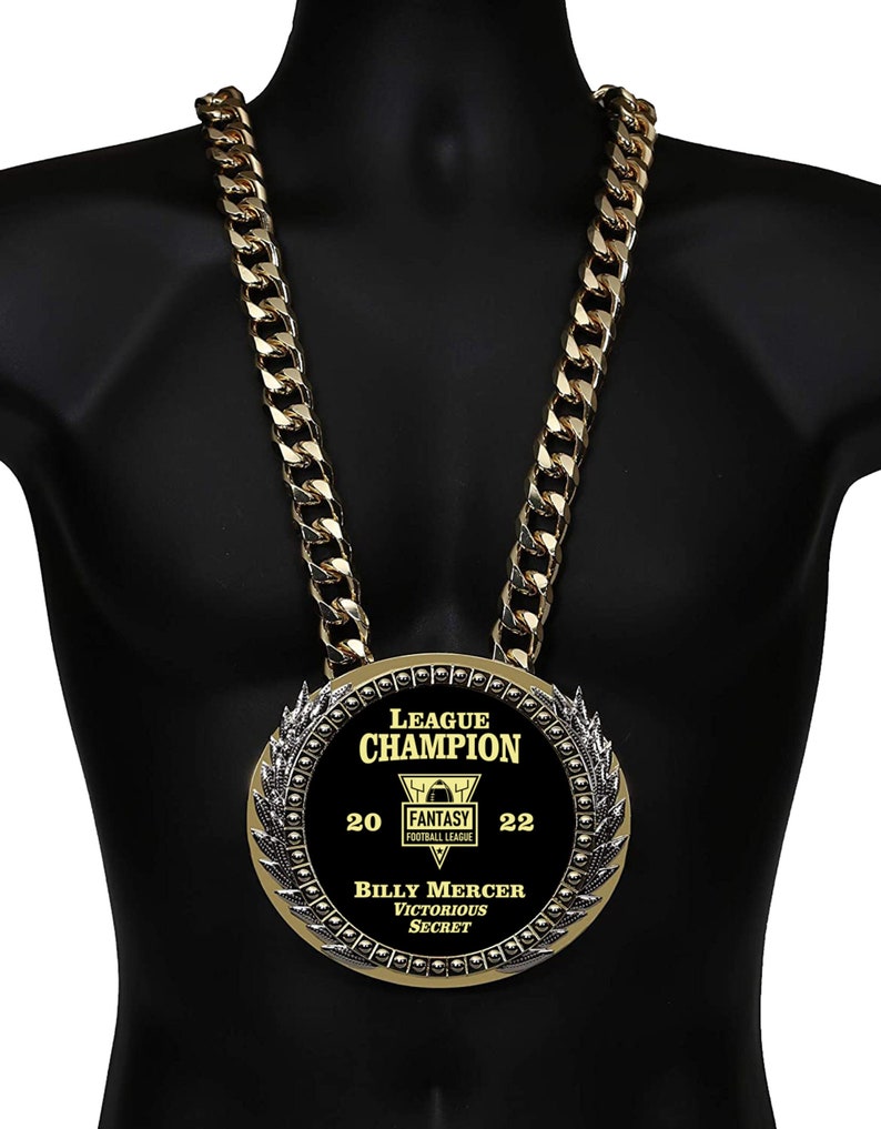 May include: A gold chain necklace with a large, round pendant. The pendant is black with gold trim and features the text "League Champion", "Fantasy Football League", "20", "22", "Billy Mercer", "Victorious Secret".