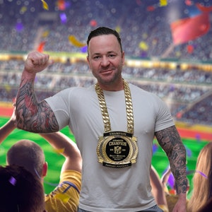May include: A man wearing a white t-shirt and a gold chain with a gold championship trophy around his neck. The trophy says "League Champion 2020" and "Billy Mercier" on it. The man is in a stadium with a crowd of people in the background.