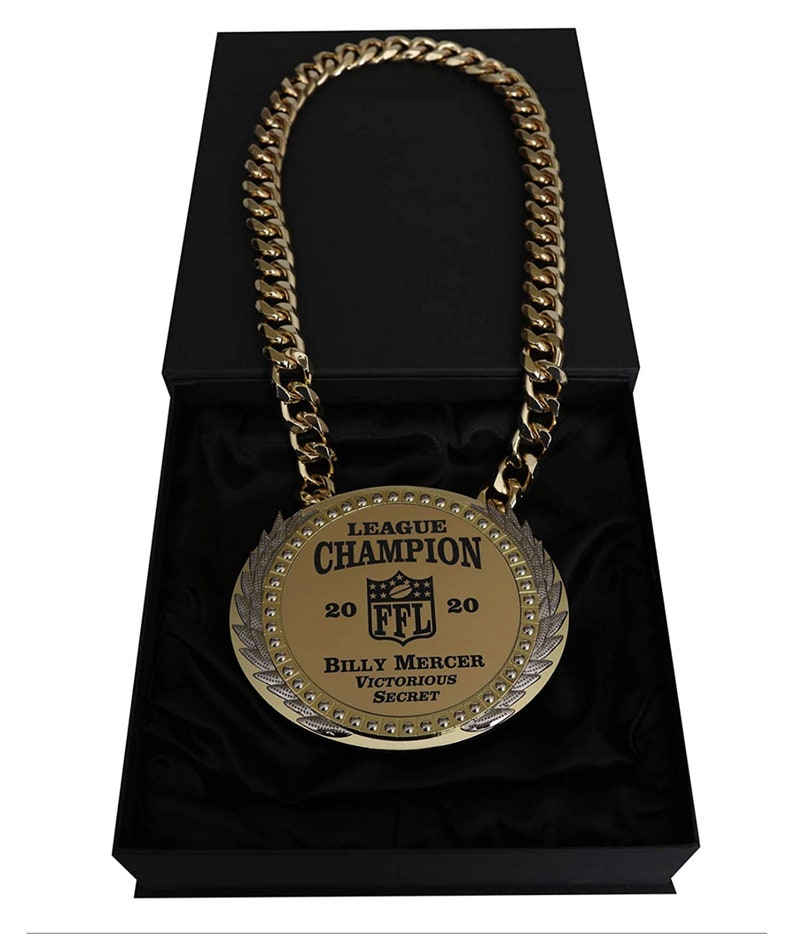 May include: A gold chain necklace with a large, round pendant. The pendant has a black and gold design with the text "League Champion 20 FFL 20 Billy Mercer Victorious Secret".