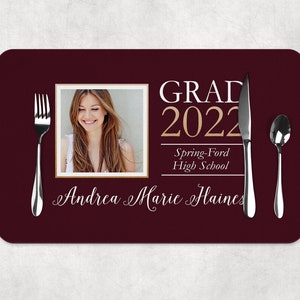 May include: A maroon placemat with a gold and white border. The placemat features a photo of a woman, the text "GRAD 2022", "Spring-Ford High School", and the name "Andrea Marie Haines".