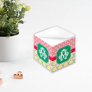 Könnte beinhalten: Ein Notizblockhalter mit rosa und grünem Muster und einem weißen Monogramm in einem grünen Kreis. Der Halter ist mit weißem Notizblockpapier gefüllt.