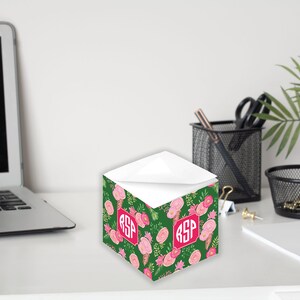 Könnte beinhalten: Ein grüner und rosa floral gemusterter Notizblockhalter mit dem Monogramm "RSP" in einem roten Quadrat. Der Halter steht auf einem weißen Schreibtisch mit einem Laptop, einem schwarzen Netzstiftbehälter und einem weißen Notizblock.