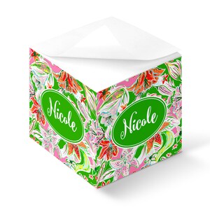 Könnte beinhalten: Ein grüner und rosa floral bedruckter Notizblock mit dem Namen "Nicole" in weiß auf einem grünen Oval. Der Notizblock hat die Form eines Würfels und eine weiße Oberseite.