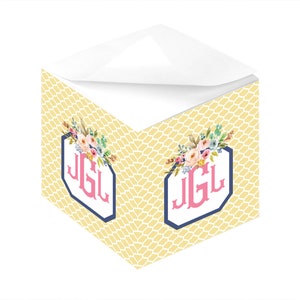 Könnte beinhalten: Ein gelber und weißer Notizblock mit einem Blumendesign und dem Monogramm "JGL" in Rosa. Der Notizblock hat die Form eines Würfels und eine weiße Oberseite.