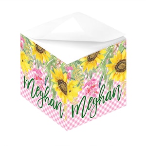 May include: A pink and white gingham notepad with a floral design featuring sunflowers and greenery. The notepad has the name "Meghan" written in green cursive on the front.