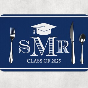 Puede incluir: Salvamanteles azul con ribete blanco, con un gorro de graduación, las letras "SMR" con rayas y el texto "CLASS OF 2025". Un tenedor, un cuchillo y una cuchara están colocados en el salvamanteles.