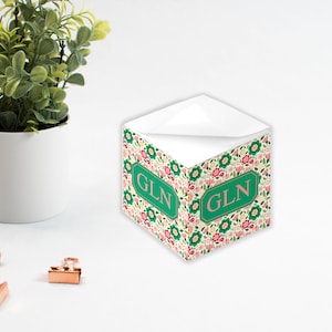 Könnte beinhalten: Ein weißer, würfelförmiger Notizblock mit einem Blumenmuster und den Initialen "GLN" in einem grünen Rechteck auf jeder Seite. Der Notizblock steht auf einer weißen Oberfläche.