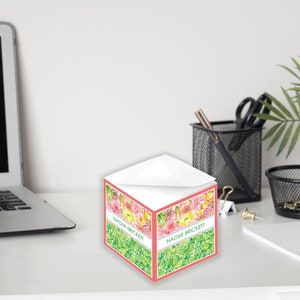May include: A pink and green floral patterned notepad with the name "Naomi Brickett" printed on the front. The notepad is sitting on a white desk.