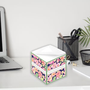 Könnte beinhalten: Ein weißer, würfelförmiger Notizblock mit einem Blumendesign. Das Design zeigt rosa, orange und gelbe Blumen auf einem dunkelblauen Hintergrund. Der Text "WELLINGTON FAMILY" ist auf dem Notizblock gedruckt.