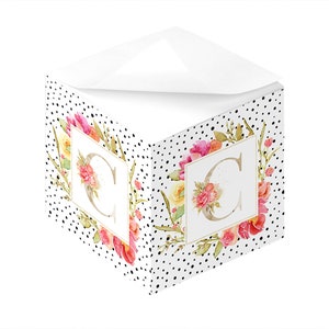 Könnte beinhalten: Ein weißer, würfelförmiger Notizblock mit einem schwarz-weißen Punkte-Muster. Die Vorder- und Rückseite des Notizblocks zeigen einen Blumenkranz mit einem goldenen Buchstaben "C" in der Mitte.