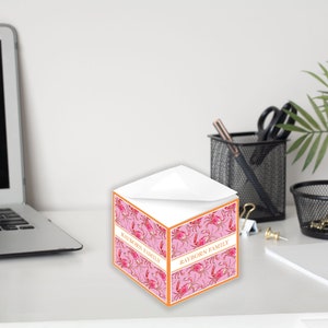 Könnte beinhalten: Ein rosa-oranger, würfelförmiger Notizblock mit einem Krabbenmuster und dem Text "Rawborn Family" auf der Vorderseite. Der Notizblock steht auf einem weißen Schreibtisch.