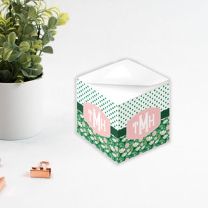 May include: A green and white floral patterned notepad holder with pink polka dots and the monogram "TMH" in a pink oval. The holder is sitting on a white surface.