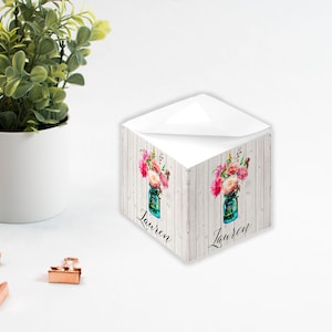 Könnte beinhalten: Ein weißer, würfelförmiger Notizblock mit einem floralen Design und dem Namen "Lauren" darauf gedruckt. Das Design zeigt ein blaues Einmachglas, gefüllt mit rosa und weißen Blumen auf einem weißen Holzgrund.