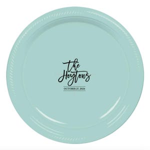 May include: A round, light blue plate with a textured rim. The center of the plate features the words "The Hoytons" in a cursive font, with the date "OCTOBER 22, 2024" below it in a smaller font. The plate is likely for a wedding or special event.