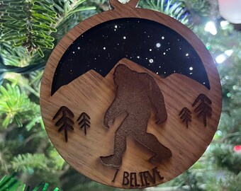 May include: A wooden Christmas ornament with a silhouette of Bigfoot walking through a snowy mountain landscape. The ornament is brown with a black background and white glitter. The text "I BELIEVE" is written at the bottom of the ornament.